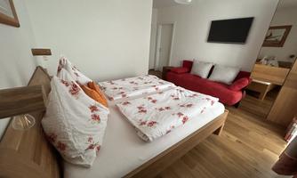 A cozy bedroom with a double bed and red cushions. In the background, there is a red sofa and a television on the wall.