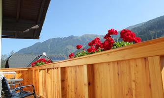 A beautiful balcony with red flowers and a view of the mountains. The sky is clear and blue.