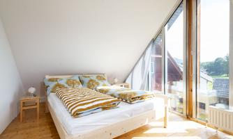 A cozy bedroom with a large window that lets in bright light. The room is minimally furnished with a wooden house bed and colorful pillows.