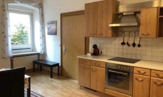 A bright kitchen with wooden cabinets and modern appliances. The room is cozy and well-structured, with a small table and a window that lets in daylight.