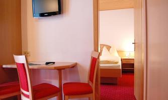 A simple hotel room with a table, two chairs, and a television on the wall. In the background, there is a bed with white pillows.