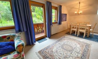 A cozy living space with a carpet, a colorful sofa, and a dining area. Large windows offer a beautiful view of the greenery.