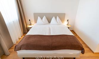 A comfortable bedroom with a large bed and modern nightstands. The bright decor and wooden flooring create an inviting atmosphere.