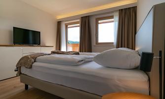 A modern bedroom with a large bed and soft pillows. The room has large windows, curtains, and a television on the wall.