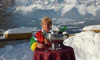 A woman is sitting at a table in the snow and enjoying the view of snow-covered mountains. On the table, there is a champagne cooler with drinks.
