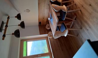 A cozy dining area with a wooden table and four chairs. The walls are partially made of wood, and there is a large window opening with a view outside.
