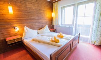 A cozy bedroom with a double bed and wooden paneling. There are large windows that let in plenty of light, and the bedding is neatly arranged.