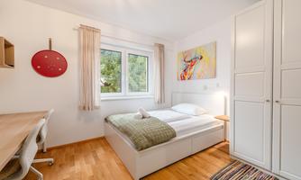 A bright bedroom with a bed, a desk, and a wardrobe. A colorful painting hangs on the wall, and there are large windows with a view of the greenery.