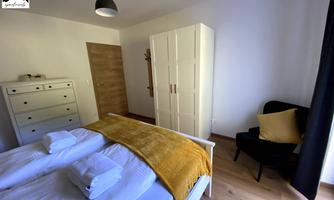 A modern bedroom with a double bed, pillows, and a golden blanket. The room contains a wardrobe and an armchair.