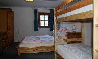 A cozy bedroom with a double bed and a bunk bed. The walls are plain, and there are wooden furniture and carpets on the floor.
