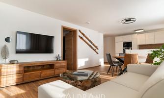 A modern living and dining room with a cozy sofa and a large television on the wall. The open kitchen is well-equipped and the space is bright and inviting.