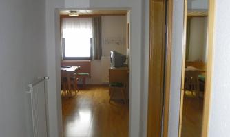 A bright hallway with wooden floors and white walls. At the end of the hallway, a window and a table are visible.