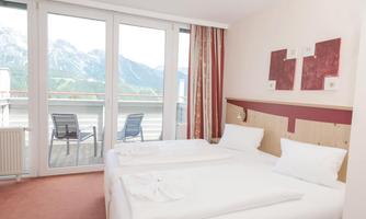 A bright, modern bedroom with two single beds. Large windows provide a view of the mountains and a balcony.