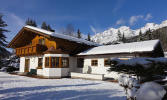A cozy house in the snow with a wooden balcony. In the background, snow-covered mountains and a blue sky are visible.