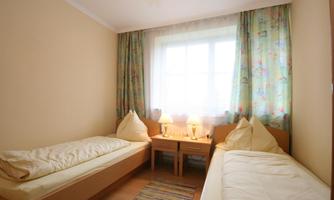 A simple bedroom with two single beds and a table in the middle. The room has yellow walls and colorful curtains that let in plenty of daylight.