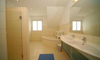 A modern bathroom with a large bathtub and a double vanity. The walls are light-colored tiles, and there is a window that provides natural light.