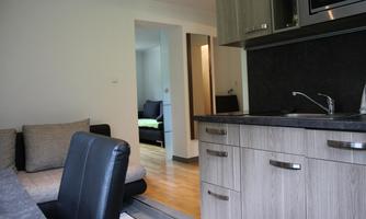 A modern kitchen with wooden cabinets and a dining area. In the background, there is a living room with a sofa.