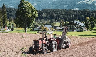 A red tractor is working a field in a rural setting. In the background, green hills and some houses are visible.