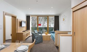 A modern living room with wooden furniture and large windows. The cozy seating area offers a view of the outdoor terrace.