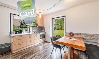 A modern kitchen with wooden cabinets and a dining table. In the background, there are windows overlooking a green garden.