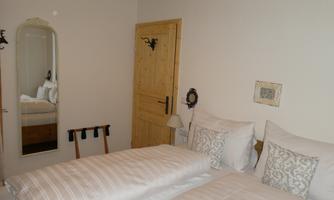 A cozy bedroom with two beds, a wooden wardrobe, and a mirror. The walls are painted light and there is a wooden door.