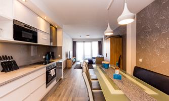 A modern kitchen with a long dining table and elegant hanging lamps. The room is bright and inviting with large windows.