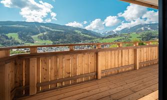 A balcony with a wooden railing offers a view of green hills and the mountains in the background. The sky is blue with some clouds.