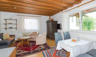 A cozy living space with wooden beam ceiling and a large window. The room is decorated with a sofa, a table, and rugs.