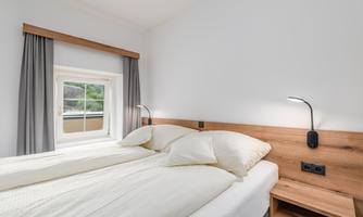 A modern bedroom with a double bed and a window. The walls are painted light, and there is a wooden bedside table.