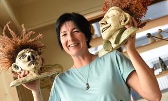 A woman is holding two mask-like sculptures with creative hairstyles in her hands. In the background, a bright room is visible.