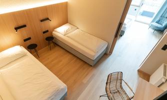 A modern room with two beds and minimalist decor. The room features a beautiful wooden floor and plenty of daylight.