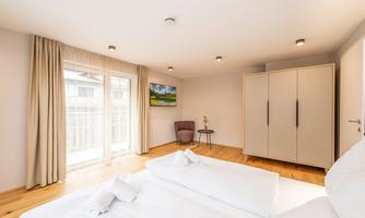 A modern bedroom with a large window and wooden floor. There is a wardrobe, a television, and a cozy armchair.
