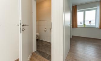 A bright room with wooden floors and large windows. In the foreground, an open door to a bathroom is visible.