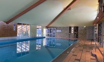 An indoor pool with clear water and a bright, friendly atmosphere. The ceiling is decorated with wooden beams, and large windows let in plenty of light.