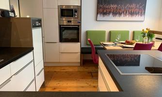 A modern kitchen with white cabinets and a cooking island. In the background, there are green chairs and a colorful piece of artwork on the wall.