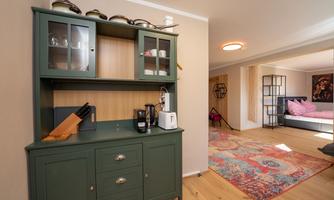 A cozy corner with a green cupboard and kitchen appliances. In the background, a bright room with a carpet and a bed is visible.