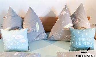 Cushions in various designs and colors are lying on a bed. The cushions have calming colors with different motifs and inscriptions.