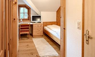 A bright hallway with wooden flooring and an open door to a cozy room. In the room, there is a bed, a desk, and a computer.