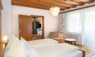 A bright and cozy room with two single beds and wooden ceilings. There is a comfortable armchair and natural light sources through large windows.