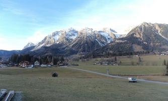 A picturesque landscape with impressive mountains in the background. In the foreground, a vast meadow stretches out with a few small buildings.