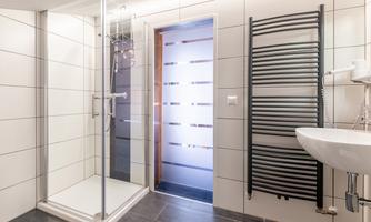 A modern bathroom with a shower, a towel radiator, and a sink. The walls are adorned with plain tiles and the lighting creates a bright atmosphere.