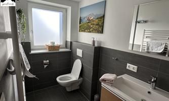 A modern bathroom with a white toilet and a wooden washbasin. A picture hangs on the wall, and there is a window that lets in a lot of light.