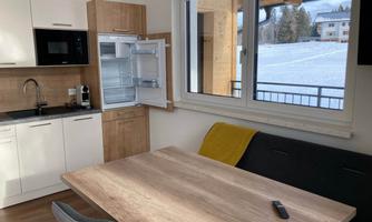 A modern kitchen with wood and white surfaces. A television is placed on the table, and through the window, one can see snow and the surrounding landscape.