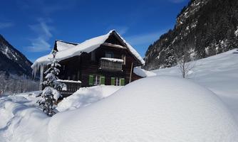 A charming wooden house in the snow, surrounded by snow-covered mountains. The sky is clear and blue, creating an idyllic winter landscape.