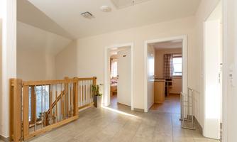 A bright hallway with a wooden railing and large windows. Through open doors, additional rooms are visible.