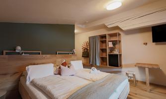 A modern room with a double bed and simple, light furniture. There is a television on the wall, and there is a cozy seating area with a plant.