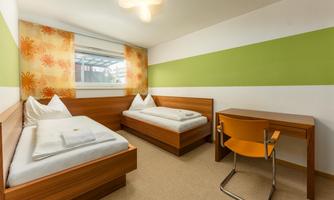 A simple room with two single beds and a desk. The walls are designed in cheerful colors, with a large window front.