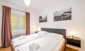 A bright bedroom with a large bed and two pillows. Black-and-white pictures hang on the walls, and there are curtains as well as a window with plenty of daylight.