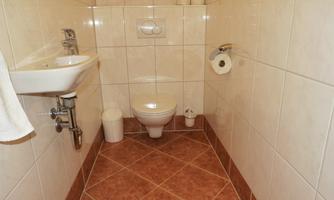 A simple bathroom with white tiles and a brown floor. It is equipped with a sink, a toilet, and toilet paper.