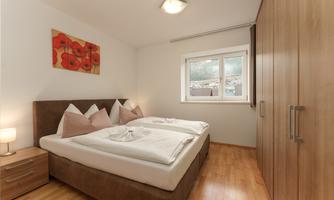 A bright bedroom with a double bed and colorful pillows. Next to the bed is a nightstand with a lamp, and a large wardrobe is present.